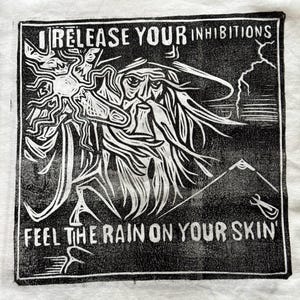 May include: A black and white linocut print on fabric featuring a bearded figure with a hat, holding a glowing orb. The text reads 'RELEASE YOUR INHIBITIONS' at the top and 'FEEL THE RAIN ON YOUR SKIN' at the bottom, with a mountain and lightning.