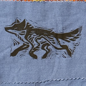 May include: A blue fabric square with a dark brown linocut print of a running wolf. The wolf is depicted in a stylized, flowing design. The fabric has a visible weave and a decorative stitched edge.