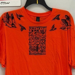 May include: Vibrant orange t-shirt with a black graphic. The design includes the word "PROTECT" above an illustration of plants and animals, with "BIODIVERSITY" below. Bird illustrations are also printed on the shoulders of the tee.