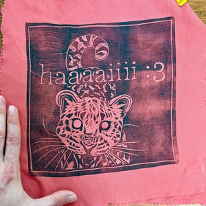 May include: Pink fabric squares with a dark red print of a leopard face and the text "haaaaiiii :3". The fabric is held together with yellow binder clips. The leopard print is inside a dark red square.