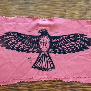 May include: A pink cloth with a black print of a bird of prey with outstretched wings. The bird is centered on the cloth, with detailed feathers and a sharp beak. The cloth has a scalloped edge.
