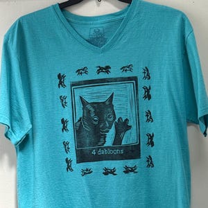 Op de afbeelding: Turquoise V-hals T-shirt met een zwarte kattenafbeelding en de tekst "4 dabloons" in een vierkant. Het shirt heeft ook een patroon van zwarte paardachtige figuren rond de kattenafbeelding.