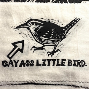以下が含まれることがあります： オフホワイトの長方形の布製パッチで、小さな鳥の黒いリノカット風イラストが描かれています。鳥を指す矢印があり、イラストの下には太いブロック体の黒い文字で「GAY ASS LITTLE BIRD」と印刷されています。
