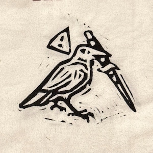 May include: A black ink block print graphic on a light beige fabric surface. The design features a stylised bird holding a dagger in its beak, positioned below a triangular warning symbol containing an exclamation point, rendered in a bold, textured style.