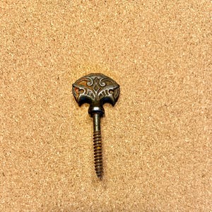 May include: An antique metal screw with an ornate, decorative head. The head features a detailed, symmetrical design with a slightly rusted appearance. The screw is a dark, aged metal, likely for furniture or hardware.