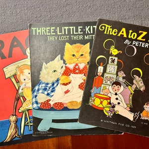 May include: Three vintage children's books with colorful illustrations. The first book is red with the word "RA" on the cover. The second book is black with the title "THREE LITTLE-KIT THEY LOST THEIR MITT" and a picture of three kittens. The third book is black with the title "The A to Z Book By PETER MABIE" and a picture of a clown and other toys.