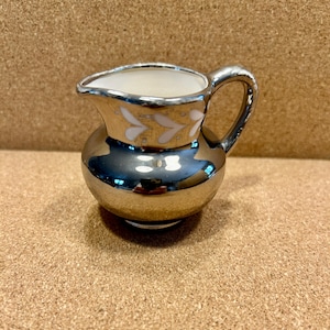 May include: A small, silver-colored creamer with a rounded body and a handle. The rim and handle are silver, while the body has a reflective, mirrored finish. White leaf-shaped designs adorn the upper portion of the creamer. The interior is white.