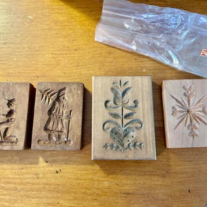 May include: Four rectangular wooden cookie molds featuring carved designs. The first shows a person sitting, the second a person carrying a tree, the third a floral heart motif, and the fourth a star-like pattern. Each mold is made of light brown wood.