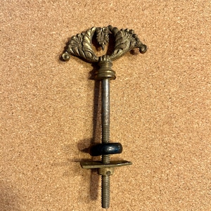 May include: An antique brass-toned furniture handle with a decorative leaf design. The handle is attached to a threaded rod with a black washer and a square metal plate. The handle is likely for a drawer or cabinet.