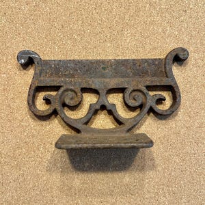 May include: A weathered, cast iron shelf bracket with an ornate, scrollwork design. The bracket has a small shelf and decorative curves at the top. The metal has a rusty, aged appearance.