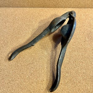 May include: A vintage, cast-iron citrus juicer with a dark, aged finish. The juicer features a hinged design with two curved handles and a bowl-shaped pressing area. The tool is designed to extract juice from citrus fruits.