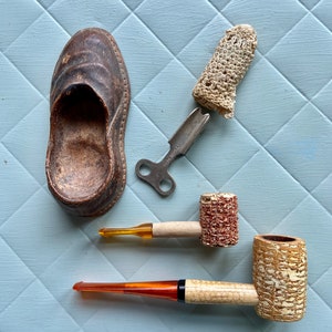 May include: A collection of vintage items including a leather slipper, a crocheted finger puppet, a metal key, and two corncob pipes with amber stems.