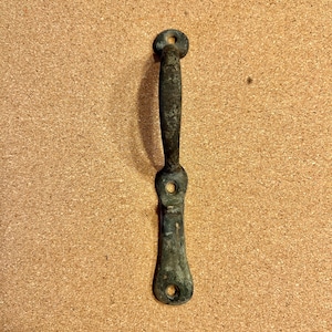 May include: A weathered, antique metal handle with a dark, aged patina. The handle features a circular top with a hole, a textured grip, and a flared base with a hole. The hardware is likely for a cabinet or drawer.