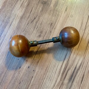 May include: A wooden door knob with two round knobs connected by a metal rod. The knobs are a light brown color and the rod is a dark metal color.