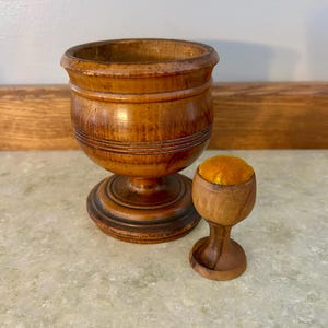 May include: A dark brown wooden darning egg holder with a pedestal base and a smaller matching wooden darning tool featuring a bright yellow fabric cushion top. Both items have a smooth, polished finish with visible wood grain and circular grooves.