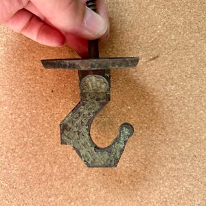 May include: A vintage-style metal hook with a screw. The hook has a decorative, textured design with a tarnished, aged appearance. The hook is a dark, muted green color.