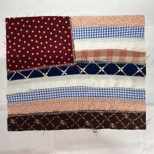 May include: A rectangular fabric wall hanging featuring a patchwork design resembling an American flag. It includes a dark red section with white stars, alternating horizontal strips of floral, gingham, solid white, and patterned fabric with frayed edges.
