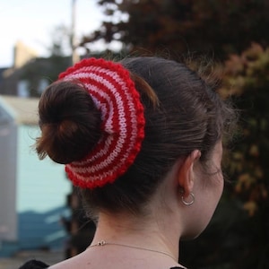 May include: A red and white striped knitted hair accessory encircles a bun hairstyle. The circular design features alternating bands of red and white, creating a bold, eye-catching pattern. The accessory adds a pop of colour and texture to the hairstyle.