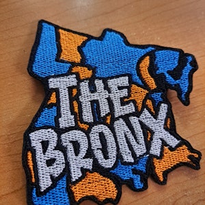 May include: Embroidered patch in the shape of The Bronx, New York. The patch features a blue and orange design with the words "THE BRONX" in white block letters. The patch has a black border.