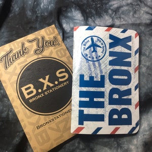 May include: Two notebooks, one with a black circle and the text "B.X.S. BRONX STATIONERY" and the other with a blue and white design with the text "THE BRONX" and a small airplane graphic.