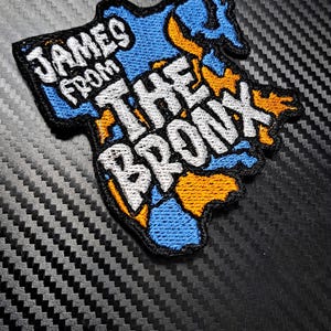 May include: A blue and orange embroidered patch with the text "James from the Bronx" in white lettering. The patch is shaped like the state of New York.