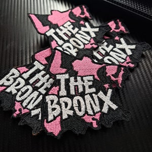 May include: Three black and pink embroidered patches shaped like the outline of the Bronx borough in New York City. Each patch says "The Bronx" in white lettering.