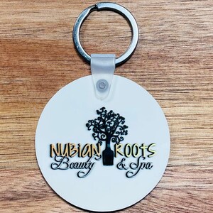 May include: White round keychain with a black tree and gold text that reads "Nubian Roots Beauty & Spa".