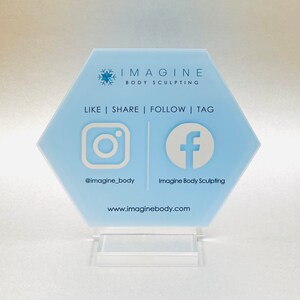 May include: A light blue hexagonal sign with the text "IMAGINE BODY SCULPTING" and a snowflake logo. The sign also includes the text "LIKE | SHARE | FOLLOW | TAG" and social media icons for Instagram and Facebook with the handles "@imagine_body" and "Imagine Body Sculpting" and the website "www.imaginebody.com".