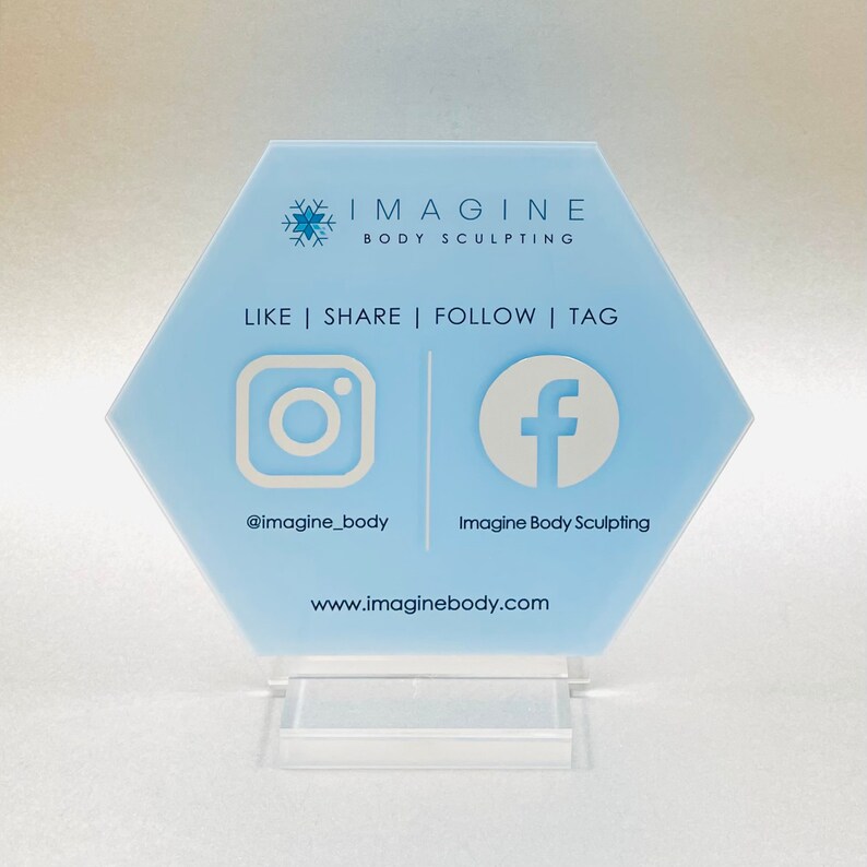May include: A light blue hexagonal sign with the text "IMAGINE BODY SCULPTING" and a snowflake logo. The sign also includes the text "LIKE | SHARE | FOLLOW | TAG" and social media icons for Instagram and Facebook with the handles "@imagine_body" and "Imagine Body Sculpting" and the website "www.imaginebody.com".