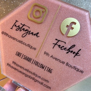May include: A pink hexagonal sign with gold glitter and gold accents. The sign has the Instagram and Facebook logos and the text "Instagram @9thavenueboutique" and "Facebook 9th Avenue Boutique" with the tagline "LIKE | SHARE | FOLLOW | TAG" and contact information.