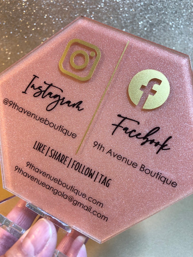 May include: A pink hexagonal sign with gold glitter and gold accents. The sign has the Instagram and Facebook logos and the text "Instagram @9thavenueboutique" and "Facebook 9th Avenue Boutique" with the tagline "LIKE | SHARE | FOLLOW | TAG" and contact information.