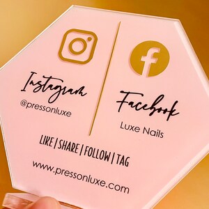 May include: Pink hexagonal sign with gold Instagram and Facebook logos. The text reads "Instagram @pressonluxe" and "Facebook Luxe Nails". The sign also includes the text "LIKE | SHARE | FOLLOW | TAG www.pressonluxe.com"