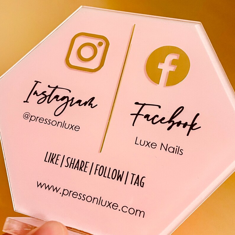 May include: Pink hexagonal sign with gold Instagram and Facebook logos. The text reads "Instagram @pressonluxe" and "Facebook Luxe Nails". The sign also includes the text "LIKE | SHARE | FOLLOW | TAG www.pressonluxe.com"