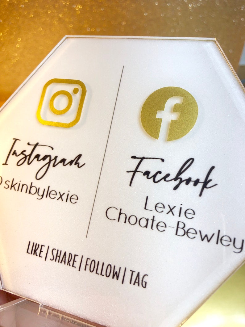 May include: White hexagonal sign with gold Instagram and Facebook logos. The text "Instagram skinbylexie" and "Facebook Lexie Choate-Bewley" is printed below the logos. The text "LIKE|SHARE|FOLLOW|TAG" is printed at the bottom of the sign.