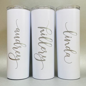 May include: Three white stainless steel tumblers with personalized names: Linda, Hillary, and Lindsey. The names are written in a silver cursive font.