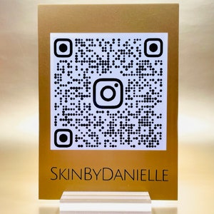 May include: A gold-colored card with a black and white QR code featuring the Instagram logo. The text "SKINBYDANIELLE" is printed below the QR code.