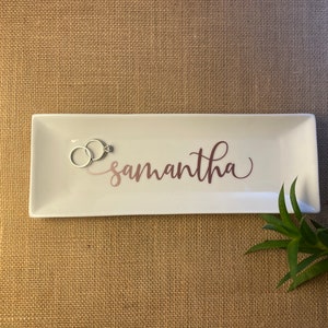 May include: White rectangular ceramic dish with a rose gold inscription that reads "samantha". Two silver rings are placed on the dish.