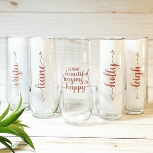 May include: Five clear glass champagne flutes with rose gold lettering. The flutes are arranged in a row on a white wooden surface. The lettering on the flutes reads "crista", "lianne", "so many beautiful reasons to be happy", "holly", and "leigh".