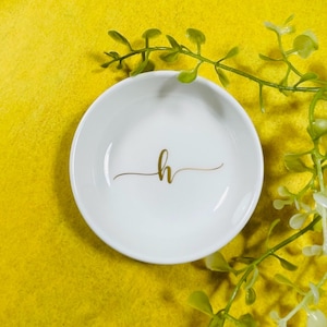 May include: A small, white ceramic dish with a gold cursive letter "h" in the center. The dish is round with a slightly raised edge. The background is a bright yellow, and there are green leafy sprigs in the background.