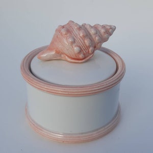 May include: A white ceramic trinket box with a pink seashell lid. The box has a pink rim and is decorated with a pink seashell.