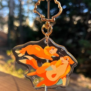 May include: A clear acrylic keychain featuring two cartoon otters in shades of orange and yellow, swimming in a starry background. The keychain has a gold-colored clasp and a decorative top.