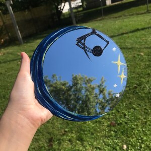May include: A round mirror with a blue and yellow painted design of a crescent moon and stars. The mirror reflects a blue sky with clouds and trees.