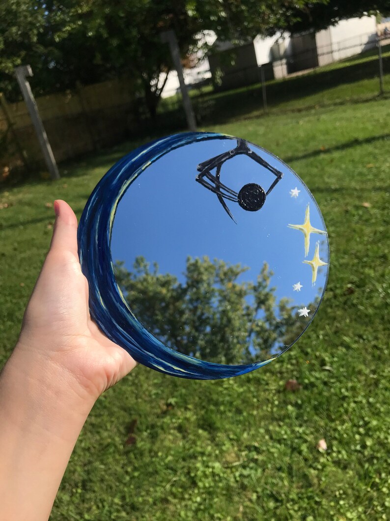 May include: A round mirror with a blue and yellow painted design of a crescent moon and stars. The mirror reflects a blue sky with clouds and trees.