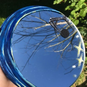 May include: A round mirror with a blue and yellow painted border, reflecting a blue sky with white stars and a black spider web.