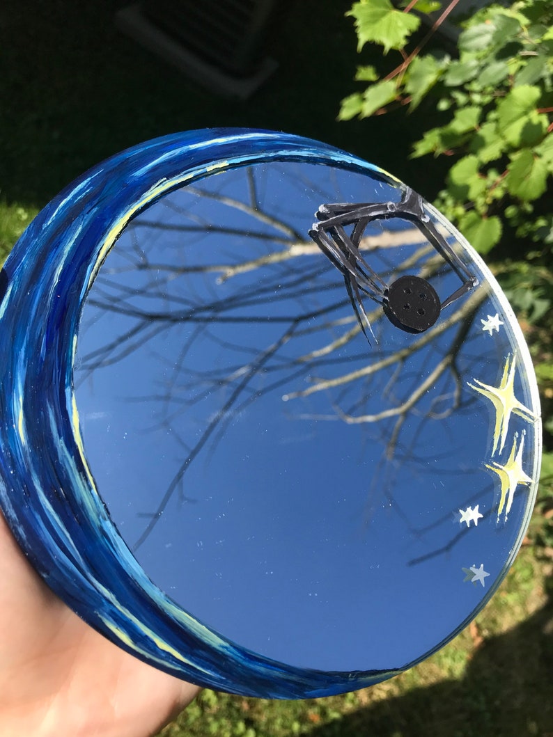 May include: A round mirror with a blue and yellow painted border, reflecting a blue sky with white stars and a black spider web.