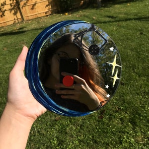 May include: A round mirror with a blue and white painted border. The mirror reflects a person holding a smartphone. The mirror has a painted design of stars and a crescent moon.