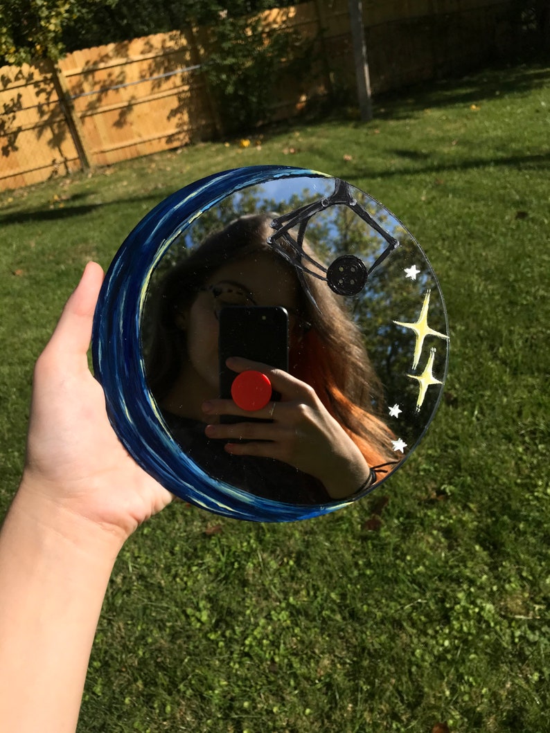 May include: A round mirror with a blue and white painted border. The mirror reflects a person holding a smartphone. The mirror has a painted design of stars and a crescent moon.