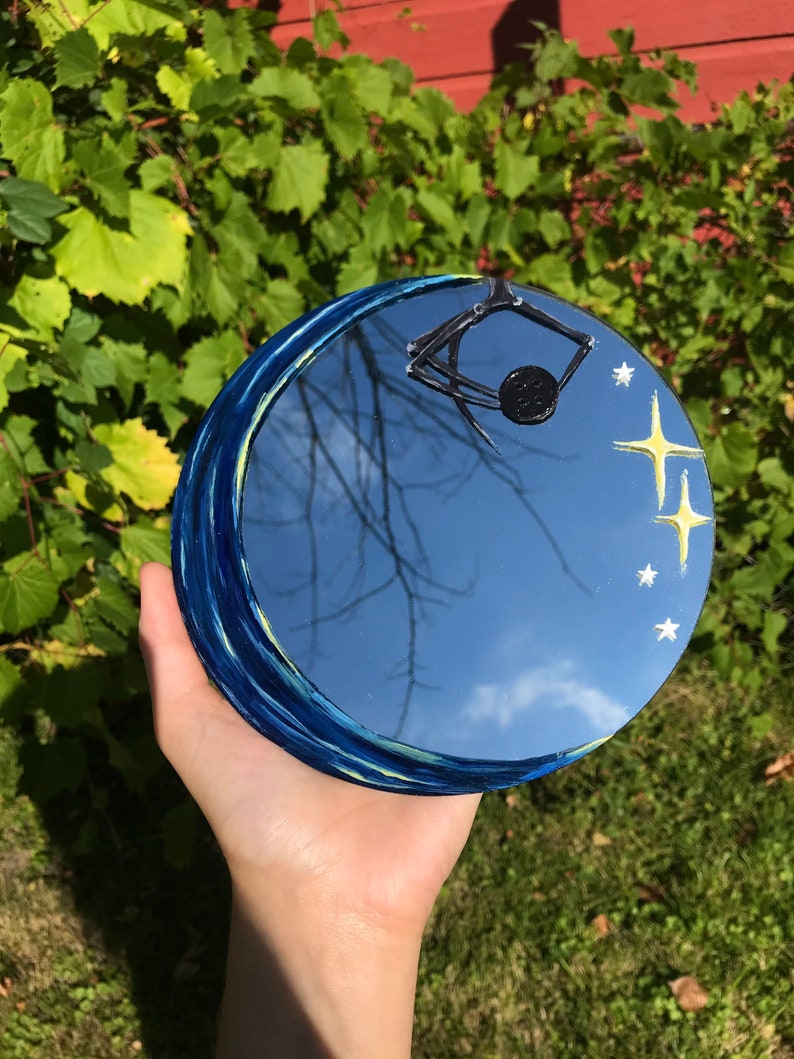 May include: A round mirror with a blue and yellow painted border depicting a starry night sky. The mirror reflects a blue sky with clouds and a tree branch. A black spider is painted on the mirror, hanging from the top.
