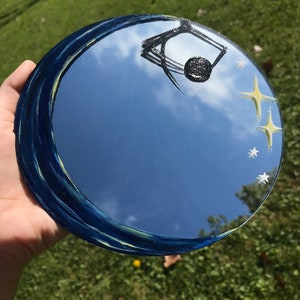 May include: A round mirror with a blue and yellow painted border. The mirror reflects a blue sky with white clouds and a black spider web design. There are yellow stars painted on the mirror.