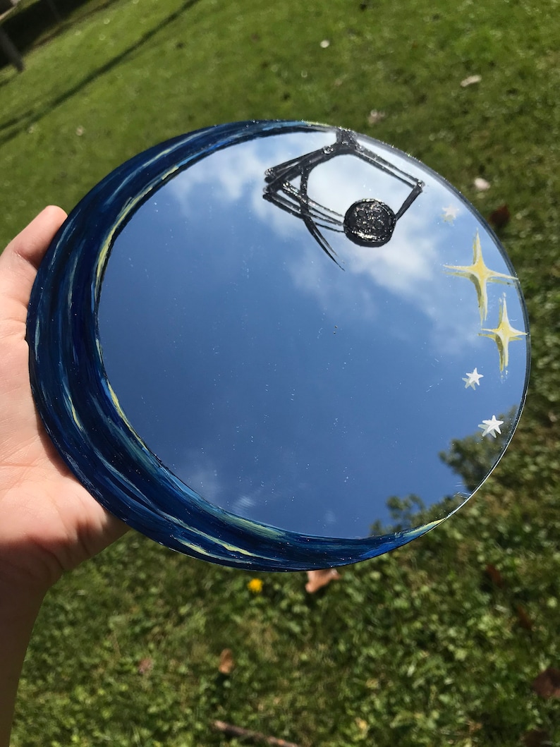 May include: A round mirror with a blue and yellow painted border. The mirror reflects a blue sky with white clouds and a black spider web design. There are yellow stars painted on the mirror.